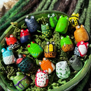 Könnte beinhalten: Eine Sammlung farbenfroher Froschfiguren in einem grünen, ovalen Behälter, der mit Moos gefüllt ist. Die Frösche sind in verschiedenen Farben bemalt, darunter Blau, Grün, Rot, Orange und Gelb, mit einzigartigen Mustern. Der Hintergrund zeigt grüne und braune dekorative Elemente.