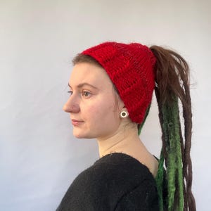 Puede incluir: Una diadema de punto roja con un agujero para la coleta. La diadema la lleva una persona con rastas largas verdes y marrones.