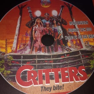 K&ouml;nnte beinhalten: Eine rot-wei&szlig;e DVD mit einem Filmplakat f&uuml;r den Film Critters. Das Plakat zeigt eine Gruppe von Menschen, die vor einem Holzzaun stehen, mit einem rot-wei&szlig;en Schild mit der Aufschrift "Critters They Bite!" Das Plakat enth&auml;lt auch den Text "MONSTERVISION FEBRUARY 12TH 2000 HOSTED BY JOE BOB BRIGGS".