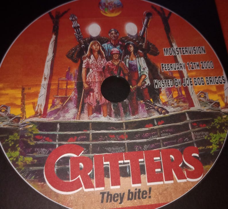 K&ouml;nnte beinhalten: Eine rot-wei&szlig;e DVD mit einem Filmplakat f&uuml;r den Film Critters. Das Plakat zeigt eine Gruppe von Menschen, die vor einem Holzzaun stehen, mit einem rot-wei&szlig;en Schild mit der Aufschrift "Critters They Bite!" Das Plakat enth&auml;lt auch den Text "MONSTERVISION FEBRUARY 12TH 2000 HOSTED BY JOE BOB BRIGGS".