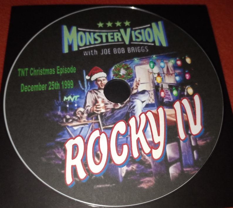 K&ouml;nnte beinhalten: Eine DVD-Disc mit schwarzem Hintergrund und einem bunten Bild eines Mannes mit Weihnachtsm&uuml;tze, der in einem Wohnmobil sitzt. Der Text "MonsterVision with Joe Bob Briggs" befindet sich oben auf der Disc. Der Text "TNT Christmas Episode December 25th 1999" befindet sich in der oberen linken Ecke. Der Text "Rocky IV" befindet sich in gro&szlig;en roten Buchstaben in der unteren rechten Ecke.