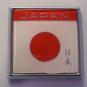 May include: A silver-framed emblem with a white background and a red circle in the center. The words "JAPAN" are printed in silver at the top of the emblem. The Japanese word for "Japan" is printed in silver at the bottom right of the emblem.