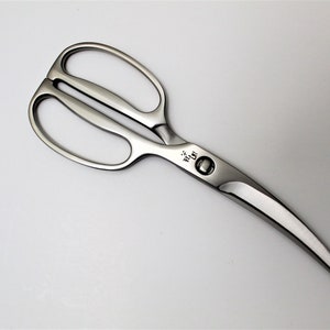 Może przedstawiać: Srebrne nożyczki kuchenne z zakrzywionym ostrzem i małym okrągłym przyciskiem na uchwycie. Nożyczki są oznaczone "Wüsthof" na uchwycie.