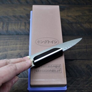 May include: A close-up of a brown sharpening stone with a purple base. The stone has Japanese text that reads "King Stone #800 Coarse #6000 Fine". A hand is holding a knife against the stone.
