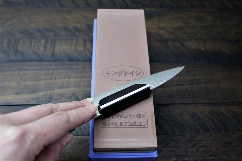 May include: A close-up of a brown sharpening stone with a purple base. The stone has Japanese text that reads "King Stone #800 Coarse #6000 Fine". A hand is holding a knife against the stone.