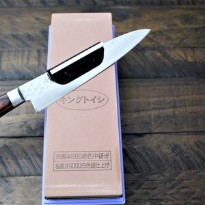 May include: A brown and purple sharpening stone with a knife resting on top. The stone has Japanese text that reads "キングトイシ" and "粒度#800赤色中研ぎ" and "粒度#6000白色超仕上げ".