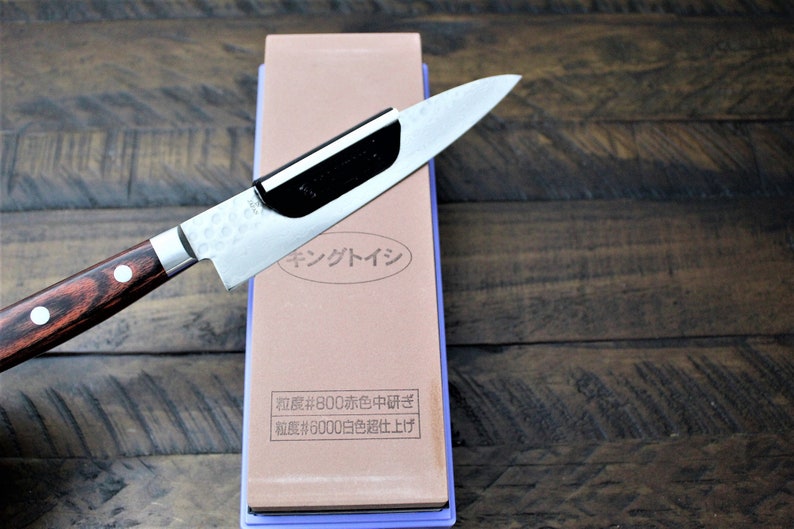 May include: A brown and purple sharpening stone with a knife resting on top. The stone has Japanese text that reads "キングトイシ" and "粒度#800赤色中研ぎ" and "粒度#6000白色超仕上げ".