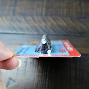 May include: A close-up of a black and silver kitchen knife with a plastic handle. The knife is packaged in a clear plastic wrapper with a blue and red label that reads "Naniwa Abrasives".