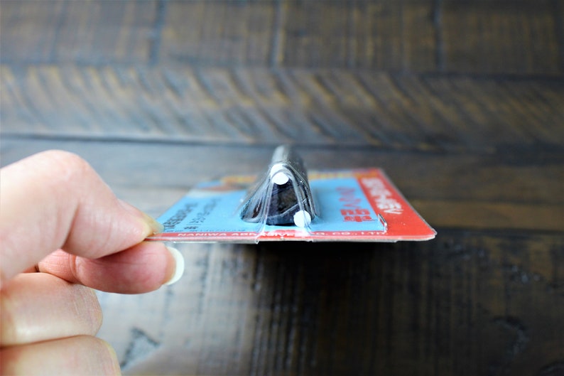 May include: A close-up of a black and silver kitchen knife with a plastic handle. The knife is packaged in a clear plastic wrapper with a blue and red label that reads "Naniwa Abrasives".