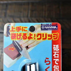 May include: A close-up of a red and white package with Japanese text that reads "NANIWA" and "上手に 研げるよ!クリップ"  The package shows a hand holding a knife and sharpening it on a red sharpening stone. The text on the package says "砥石で包" which translates to "wrapped in a whetstone".