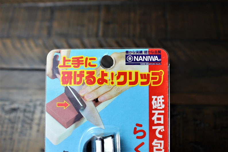 May include: A close-up of a red and white package with Japanese text that reads "NANIWA" and "上手に 研げるよ!クリップ"  The package shows a hand holding a knife and sharpening it on a red sharpening stone. The text on the package says "砥石で包" which translates to "wrapped in a whetstone".