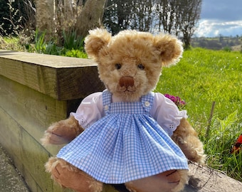 Pode incluir: Um urso de peluche castanho vestindo um vestido azul e branco xadrez com uma borda de renda branca. O urso está sentado em uma cerca de madeira com um fundo de grama verde.