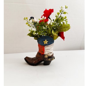 May include: A decorative cowboy boot vase filled with red, white, and green artificial flowers. The boot is painted in the colors of the Texas flag, with a blue top and a gold star. The boot is brown with gold accents.