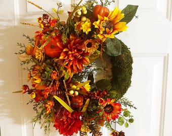 May include: A fall-themed wreath with a moss base, adorned with sunflowers, pumpkins, and various autumn foliage. The wreath features a vibrant mix of orange, yellow, and red flowers, creating a warm and inviting aesthetic. The wreath is hanging on a white door.