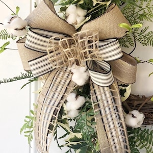 May include: A decorative wreath featuring a large burlap bow with black and white striped ribbon accents. The wreath is adorned with cotton bolls and various green foliage, creating a rustic and natural aesthetic. The bow is the focal point.
