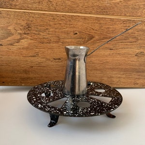 Op de afbeelding: Een zilveren koffiepot met een lange handgreep staat op een zwart-wit gespikkelde metalen standaard met drie poten.