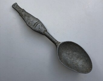 Op de afbeelding: Een zilveren metalen lepel met een unieke, getextureerde steel. De lepel heeft een ronde kom en een licht gebogen steel.