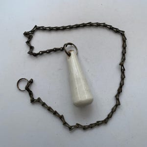 May include: A white ceramic pull handle with a smooth, tapered cylindrical shape attached to a dark, weathered metal chain. The chain features a series of interlocking rectangular links and includes a circular metal ring at one end.