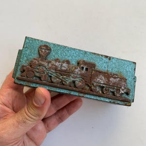 Può includere: Un timbro metallico rettangolare in stile vintage con un rilievo dettagliato di un treno a vapore. Il treno è in tonalità marroni e ruggine, su uno sfondo turchese strutturato. Il timbro è tenuto in una mano.