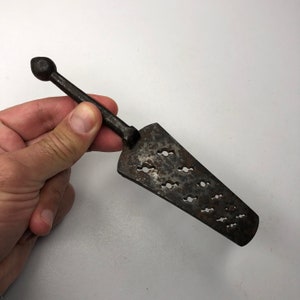 Puede incluir: Una herramienta de metal vintage con un mango puntiagudo y una hoja plana y estampada. La hoja tiene una serie de pequeños agujeros espaciados uniformemente.