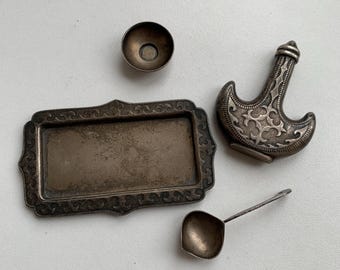 May include: A silver tray with an ornate design, a small silver bowl, a silver spoon, and a silver bottle shaped like a crescent moon.