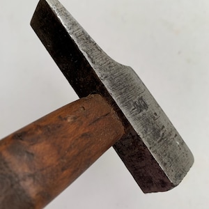 Könnte beinhalten: Ein Hammerkopf aus Metall mit einer verwitterten, dunkelgrauen Oberfläche, der an einem Holzstiel befestigt ist. Der Metallkopf verfügt über eine flache, rechteckige Schlagfläche und eine keilförmige Finne auf der gegenüberliegenden Seite.