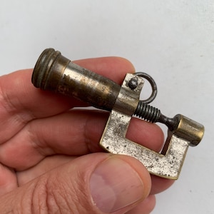 May include: A vintage brass and steel pocket micrometer featuring a C-shaped frame, a threaded spindle, and a cylindrical barrel with engraved numerical markings including the number 5. The metal surfaces show signs of age, wear, and oxidation.