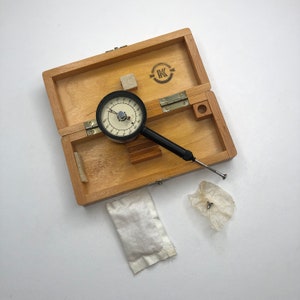 Könnte beinhalten: Eine Holzkiste mit einem Scharnierdeckel, die eine Messuhr mit einem schwarzen Griff und einem kleinen weißen Quadrat enthält. Die Kiste trägt ein Etikett mit den Buchstaben "IK". Die Messuhr dient zur Messung der Härte von Materialien.