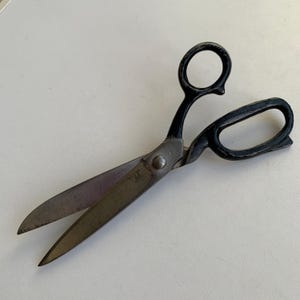 Op de afbeelding: Een zware metalen schaar met lange, puntige stalen bladen en zwart gelakte handgrepen. Het gereedschap heeft een centrale draaischroef die de twee bladen verbindt, met één ronde vingeropening en één langwerpige ovale opening.