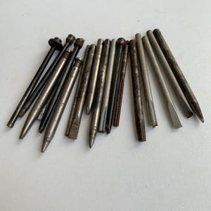 Op de afbeelding: Een verzameling van diverse metalen gereedschappen, waaronder doorslagen, drevels en beitels. Deze cilindrische instrumenten hebben een verweerde grijze en donkere stalen afwerking, met puntige uiteinden, platte koppen of taps toelopende schachten.