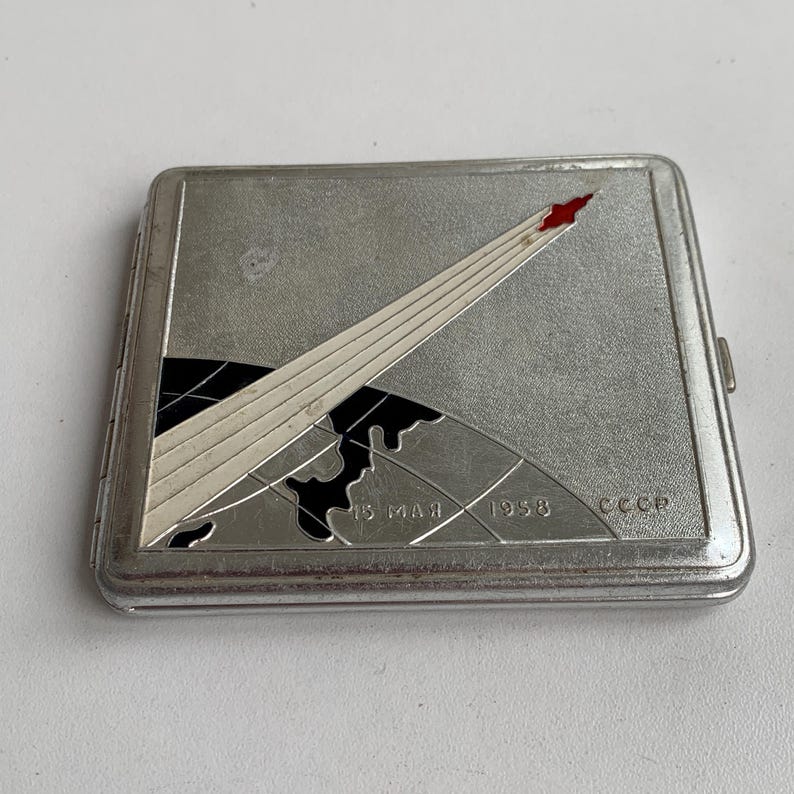 May include: A silver cigarette case featuring a rocket launching into space over a globe. The case is inscribed with the text "15 MAY 1958 CCCP" at the bottom.