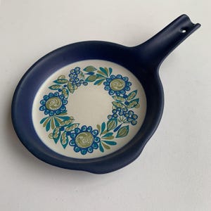 Puede incluir: Un reposacucharas de cerámica azul marino con un asa y un diseño floral. El centro del reposacucharas es blanco, con una corona de flores azules, turquesas y verdes. El asa tiene un agujero para colgar.