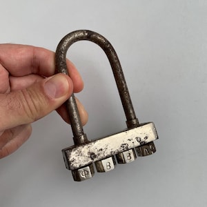Könnte beinhalten: Ein Vintage-Kombinationsschloss mit silberner Oberfläche und vier nummerierten Zifferblättern. Die Zifferblätter sind mit den Buchstaben "Φ", "B", "B" und "A" beschriftet.