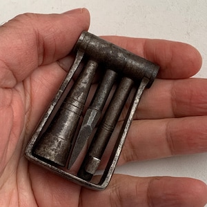 May include: A vintage metal tool set comprising three pieces within a rectangular frame. The tools have a dark, aged appearance, with one piece resembling a small blade. The set is held in a hand.