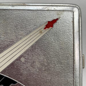 May include: A silver cigarette case featuring a red and white comet or shooting star design. The case has a textured surface and a hinge on the right side.