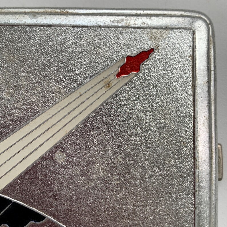 May include: A silver cigarette case featuring a red and white comet or shooting star design. The case has a textured surface and a hinge on the right side.