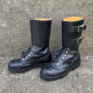 May include: A pair of black leather boots with laces and silver eyelets. The boots are mid-calf length and have two adjustable buckle straps. The soles are black and the interior is a light tan colour.