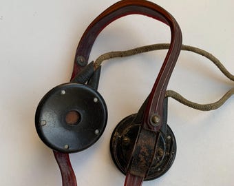 Op de afbeelding: Een paar vintage zwarte en rode koptelefoons met een stoffen snoer. De koptelefoon heeft een ronde, zwarte oortelefoon met een kleine rode cirkel in het midden.