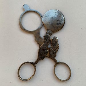 May include: Antique silver-toned scissor-like tool with a rooster design. The tool features a circular handle, a magnifying glass, and a round mirror. The rooster is intricately detailed, adding a decorative element to the functional design.