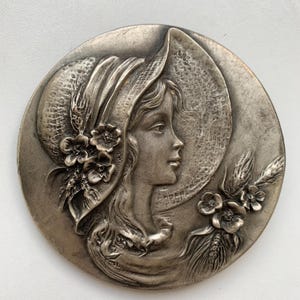May include: A circular, silver-toned relief sculpture featuring a woman in profile wearing a wide-brimmed hat adorned with flowers and wheat. The woman's face is detailed, and the overall design has a vintage aesthetic.