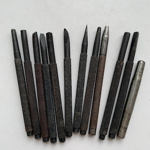 Könnte beinhalten: Eine Sammlung von zwölf Metallhandwerkzeugen, darunter verschiedene Durchschläger und Meißel mit texturierten, gerändelten Griffen. Die Werkzeuge haben dunkle, verwitterte Oberflächen und verschiedene Spitzenformen, von spitz bis flach.