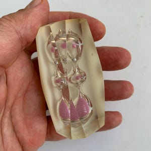 Op de afbeelding: Een helder, rechthoekig blok hars met daarin een miniatuur glazen zandloper gevuld met roze zand. De zandloper heeft een ontwerp met dubbele bollen en een smalle hals, ingebed in het doorschijnende, gebroken witte, massieve harsmateriaal.