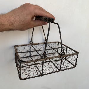 May include: A vintage wire basket with a rectangular shape and a handle made of dark wood and metal. The basket is constructed with a chicken wire mesh and has a rustic, aged appearance. The handle is attached to the basket with metal loops.
