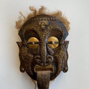 May include: A handcrafted, antique-style mask with a dark brown metal face, adorned with gold-colored accents around the eyes and forehead. The mask features a protruding metal tongue and is topped with a fringe of light brown fibers.