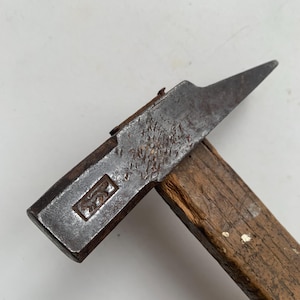 Könnte beinhalten: Ein Metallhammer mit einem verwitterten Holzgriff. Der Stahlkopf weist einen rechteckigen Stempel eines vierbeinigen Tieres innerhalb eines Rahmens auf. Die Metalloberfläche zeigt Oxidationsspuren, ein spitzes Ende und eine flache Schlagfläche.