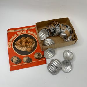 以下が含まれることがあります： クルミの形をしたペストリーを焼くためのヴィンテージメタルモールドのセット。モールドは、キリル文字で「форма для печен」と書かれた赤と白のラベルが付いた段ボール箱に入っています。ラベルには、クルミの入ったボウルも描かれています。