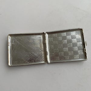 May include: Silver cigarette case with a chequered pattern on the inside and a geometric design on the outside.