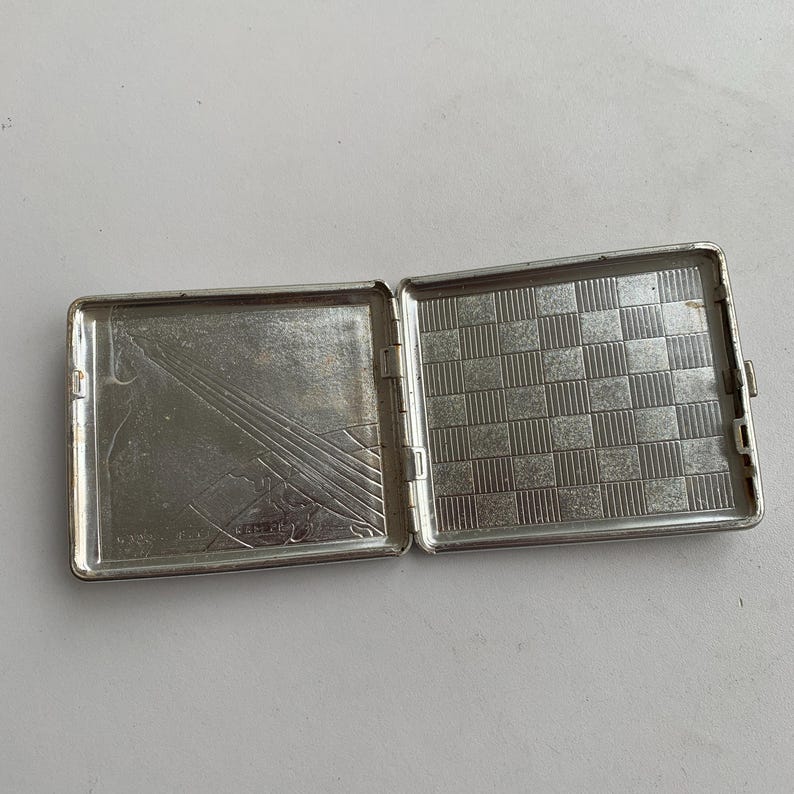 May include: Silver cigarette case with a chequered pattern on the inside and a geometric design on the outside.