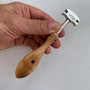 Può includere: Un tagliavetro con manico in legno chiaro, una singola vite scura e una testa in metallo argentato. La testa metallica presenta un bordo superiore curvo con la parola SPECIAL incisa in lettere maiuscole nere sul lato.
