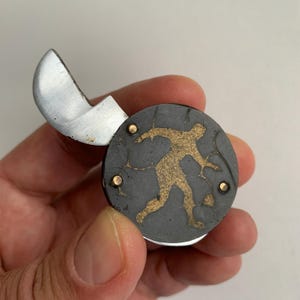 Könnte beinhalten: Ein kleines, schwarzes Taschenmesser mit einer goldenen Silhouette einer Person auf dem Griff. Das Messer hat eine silberne Klinge und einen Messingniet.